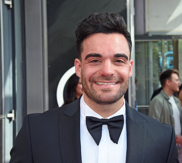 Stefano Zarrella: Ein junger Mann mit dunklen Haaren und Bart lächelt in einem schwarzen Smoking mit Fliege vor einer gläsernen Event-Location. | © IMAGO / Gartner