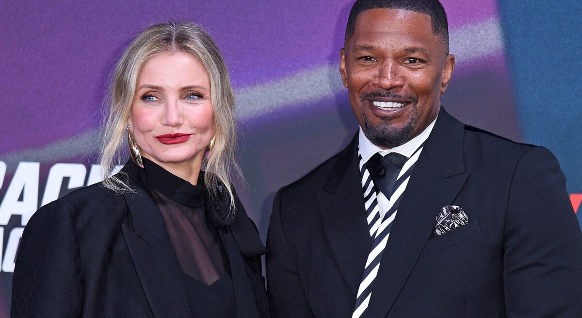 Cameron Diaz mit blonden Haaren, rotem Lippenstift und Jamie Foxx im dunklen Anzug stehen nebeneinander und lächeln vor einer bunten Wand.