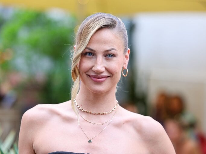 Elegante Frau in schulterfreiem Outfit, mit natürlichem Lächeln, goldenen Accessoires und unscharfem Hintergrund im Freien. | © Gerald Matzka/Getty Images
