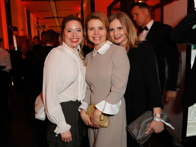 Drei Frauen in eleganter Abendgarderobe, lächelnd bei einem Event mit roter Beleuchtung und stilvollem Ambiente. | © Andreas Rentz/Getty Images