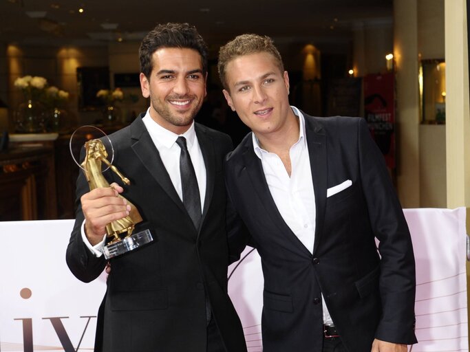 Zwei junge Männer in schwarzen Anzügen, einer hält eine goldene Trophäe, lächelnd bei einer Preisverleihung in elegantem Ambiente. | © IMAGO / Spöttel Picture