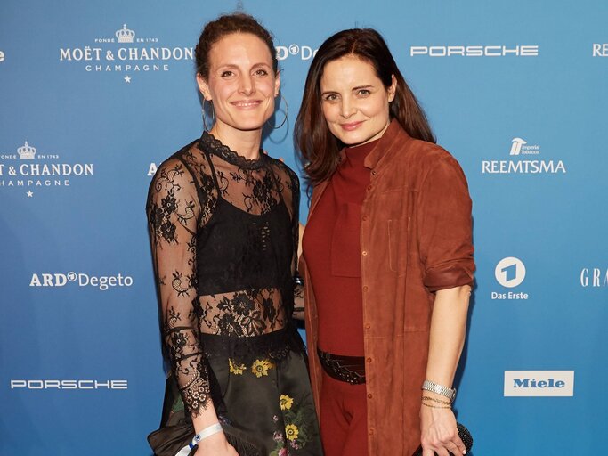 Zwei Frauen bei einem Event vor blauem Hintergrund, links in schwarzer Spitzenbluse, rechts in braunem Outfit, beide lächelnd. | © IMAGO / eventfoto54