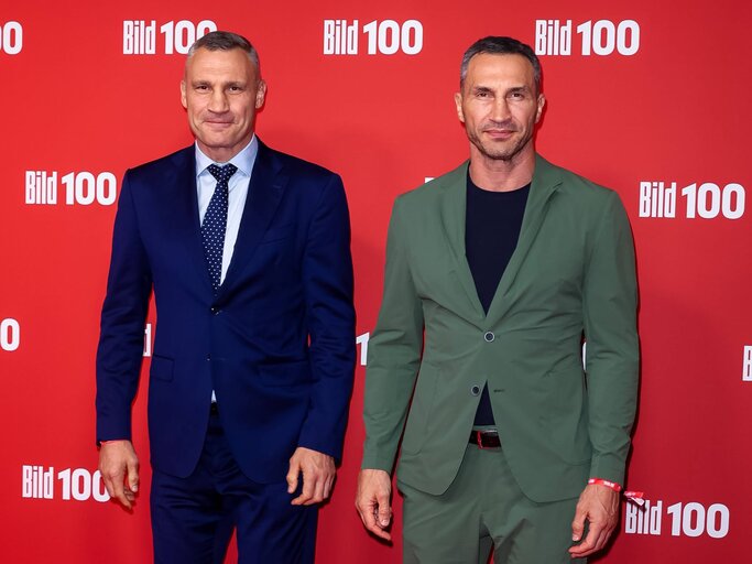 Zwei Männer posieren lächelnd in stilvoller Abendkleidung, vor einer roten Fotowand mit dem Logo „Bild 100“. | © IMAGO/ xEventpressxKochanx
