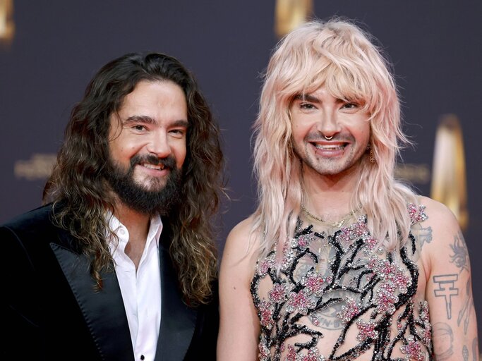 Zwei Männer mit langen Haaren, links mit Bart im schwarzen Anzug, rechts in funkelndem, transparentem Kleid mit rosa Details, beide lächelnd. | © IMAGO / Panama Pictures