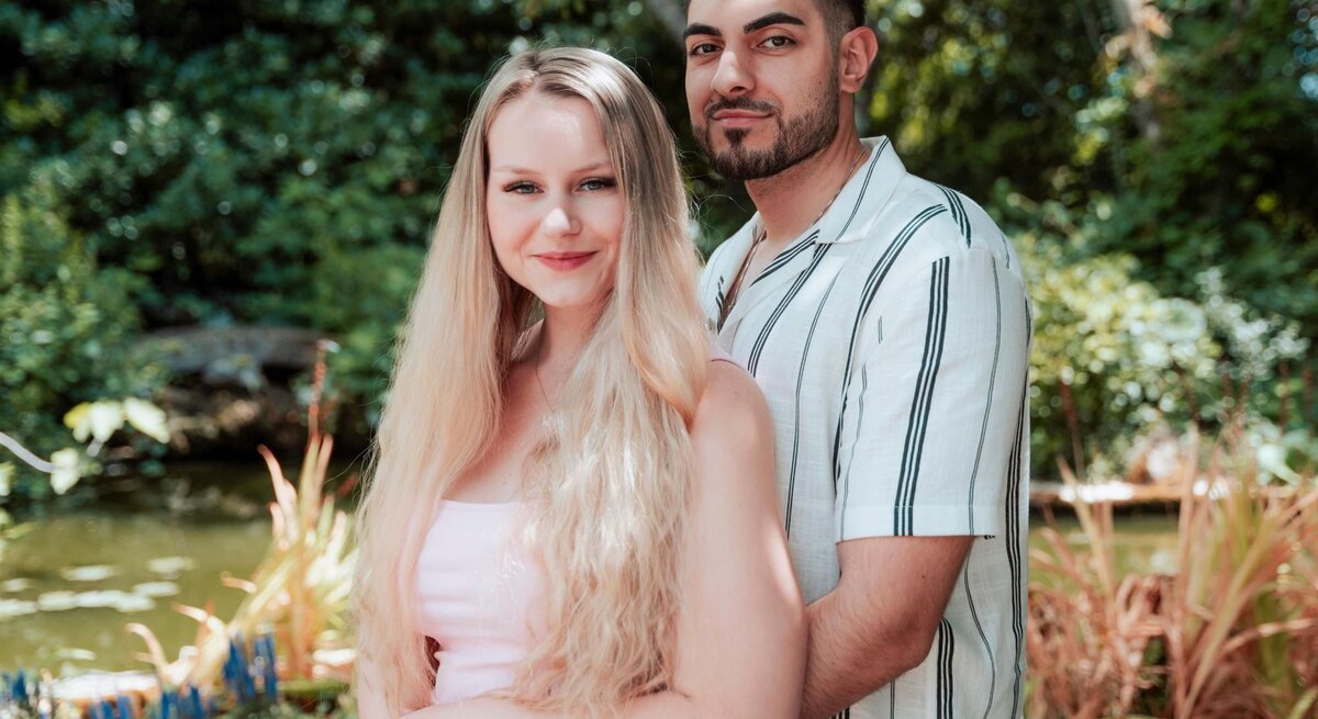 Eine blonde Frau (Sarah-Jane Wollny) und ein dunkelhaariger Mann (Tinush) posieren umarmt vor einem Teich in einem sonnigen, grünen Park.