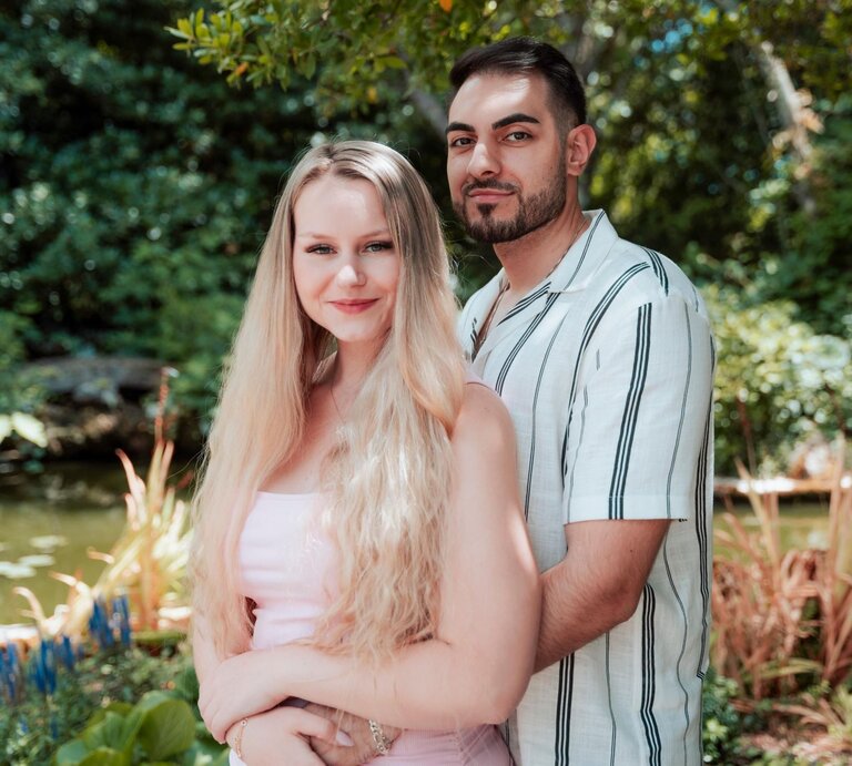 Eine blonde Frau (Sarah-Jane Wollny) und ein dunkelhaariger Mann (Tinush) posieren umarmt vor einem Teich in einem sonnigen, grünen Park. | © RTL / Kimberly Schäfer
