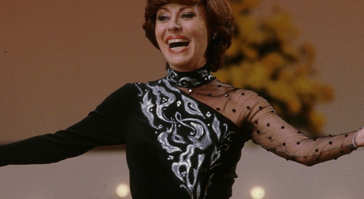 Caterina Valente auf der Bühne: Sie trägt ein elegantes schwarzes Kleid mit silbernen, dekorativen Mustern auf der Vorderseite, einem transparenten und einem gepunkteten Ärmel. Sie lacht herzlich und hat die Arme leicht geöffnet.