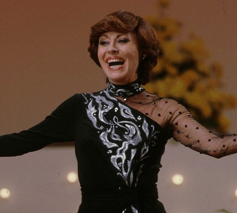 Caterina Valente auf der Bühne: Sie trägt ein elegantes schwarzes Kleid mit silbernen, dekorativen Mustern auf der Vorderseite, einem transparenten und einem gepunkteten Ärmel. Sie lacht herzlich und hat die Arme leicht geöffnet. | © Getty Images / Peter Bischoff / Freier Fotograf