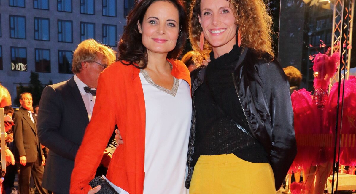 Zwei lächelnde Frauen stehen abends auf einem roten Teppich, die eine trägt ein orangefarbenes Kleid, die andere eine schwarze Jacke und gelbe Hose.