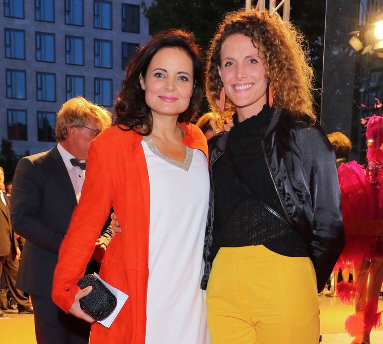 Zwei lächelnde Frauen stehen abends auf einem roten Teppich, die eine trägt ein orangefarbenes Kleid, die andere eine schwarze Jacke und gelbe Hose. | © IMAGO / Tinkeres