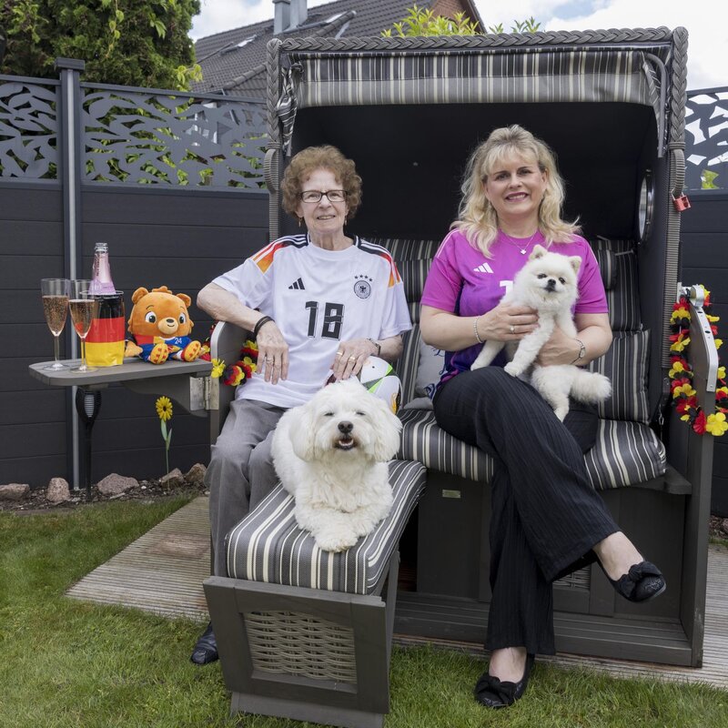 Zwei Frauen in Fußballtrikots sitzen lächelnd auf einem Strandkorb im Garten, eine hält einen weißen Hund, die andere hat einen Hund vor sich. | © Anika Büssemeier/ BILD der FRAU