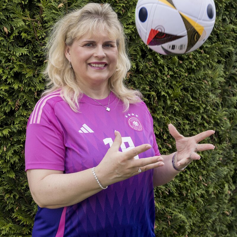 Eine Frau mit schulterlangem blondem Haar trägt ein pinkes Trikot und posiert lächelnd vor einer Deutschlandflagge. | © Anika Büssemeier/ BILD der FRAU