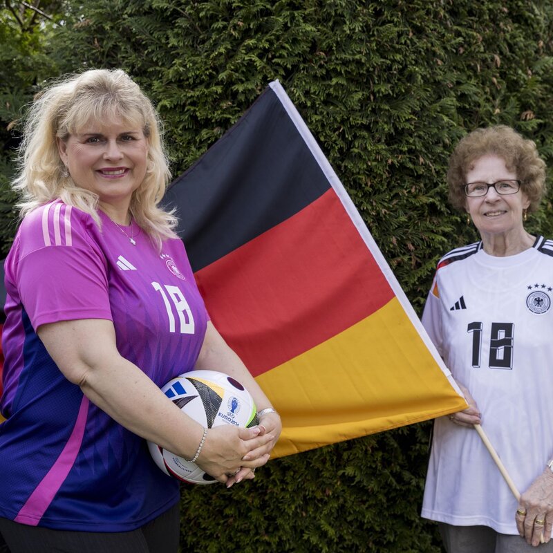 Viola Mittelstädt steht vor einer Deutschlandflagge, die Oma Angelika hält. Beide tragen ein Deutschlandtrikot und Viola hält einen Fußball in der Hand. | © Anika Büssemeier/ BILD der FRAU