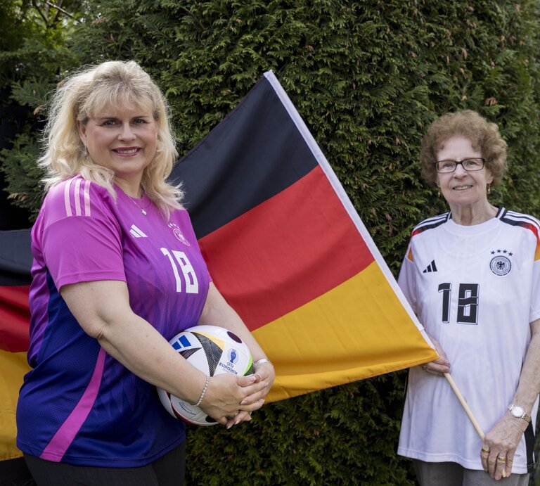 Viola Mittelstädt steht vor einer Deutschlandflagge, die Oma Angelika hält. Beide tragen ein Deutschlandtrikot und Viola hält einen Fußball in der Hand. | © Anika Büssemeier/ BILD der FRAU