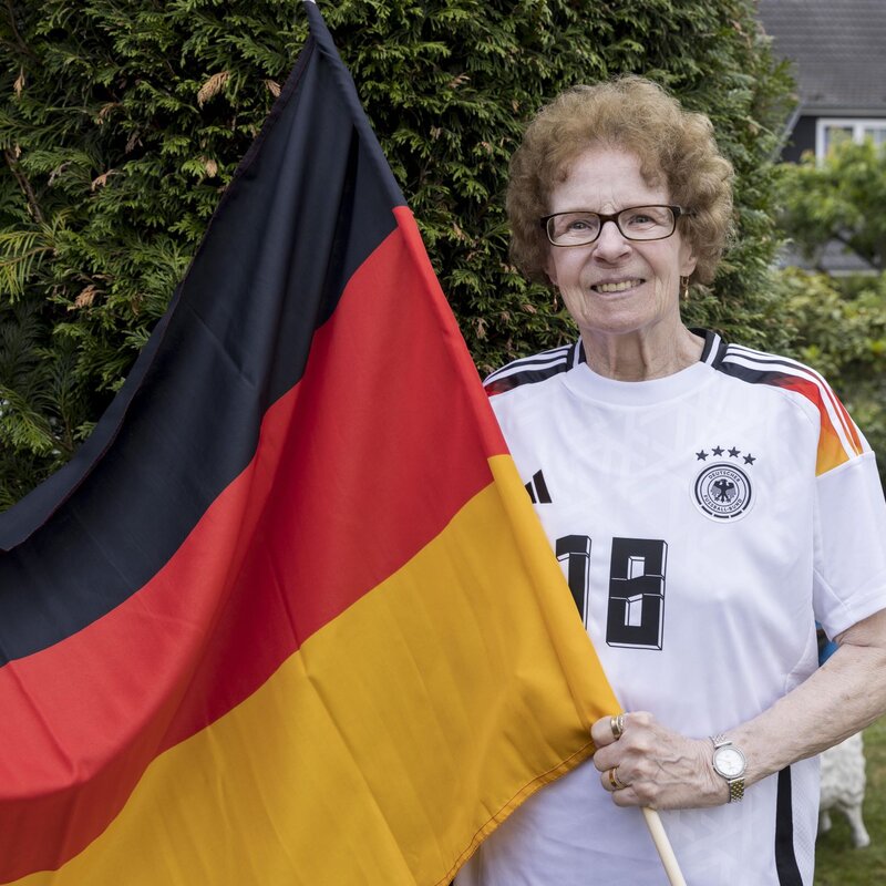 Eine ältere Frau, Oma Angelika Mittelstädt, mit lockigem Haar und Brille hält lächelnd eine große Deutschlandflagge im Garten. | © Anika Büssemeier/ BILD der FRAU