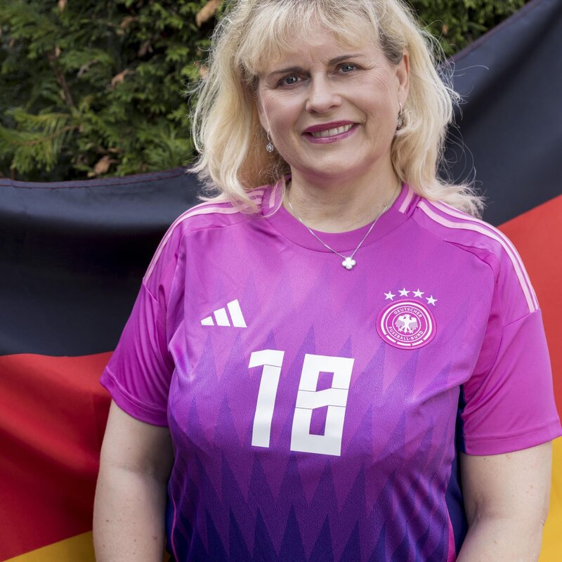 Eine lächelnde Frau im pinken Fußballtrikot der deutschen Nationalmannschaft spielt mit einem Ball vor einer dichten Hecke. | © Anika Büssemeier/ BILD der FRAU