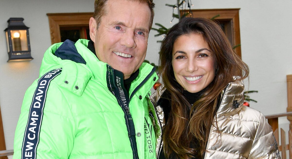 Ein Foto zeigt Dieter Bohlen und Carina Walz in Winterkleidung. Dieter Bohlen trägt eine leuchtend grüne Jacke mit dem Aufdruck "CAMP DAVID" und blickt direkt in die Kamera. Carina Walz hingegen trägt eine metallisch goldene Jacke und lächelt, während sie zur Kamera schaut. Im Hintergrund ist eine traditionelle Holzstruktur sichtbar, die auf ein alpines Setting hinweist, unterstrichen durch die winterliche Kleidung des Paares.