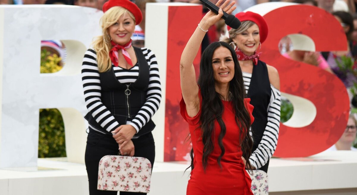 Kiki Cordalis, Tochter von Costa Cordalis, bei einem Auftritt auf einer Bühne. Sie winkt mit einem Mikro in der Hand