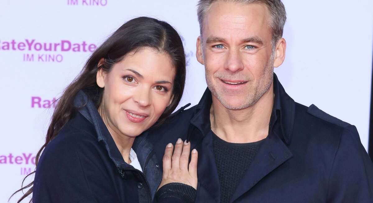 Katja Woywood und Marco Girnth  lächeln und posieren gemeinsam für ein Foto. Katja Woywood lehnt sich an Marco Girnth und schaut direkt in die Kamera, während Marco Girnth zur Seite blickt. Beide tragen dunkle Oberbekleidung. Im Hintergrund ist eine Werbetafel mit dem Schriftzug "RateYourDate IM KINO" in Pink zu sehen, was auf eine Filmpremiere oder ein ähnliches Event hinweist, bei dem sie anwesend sind.