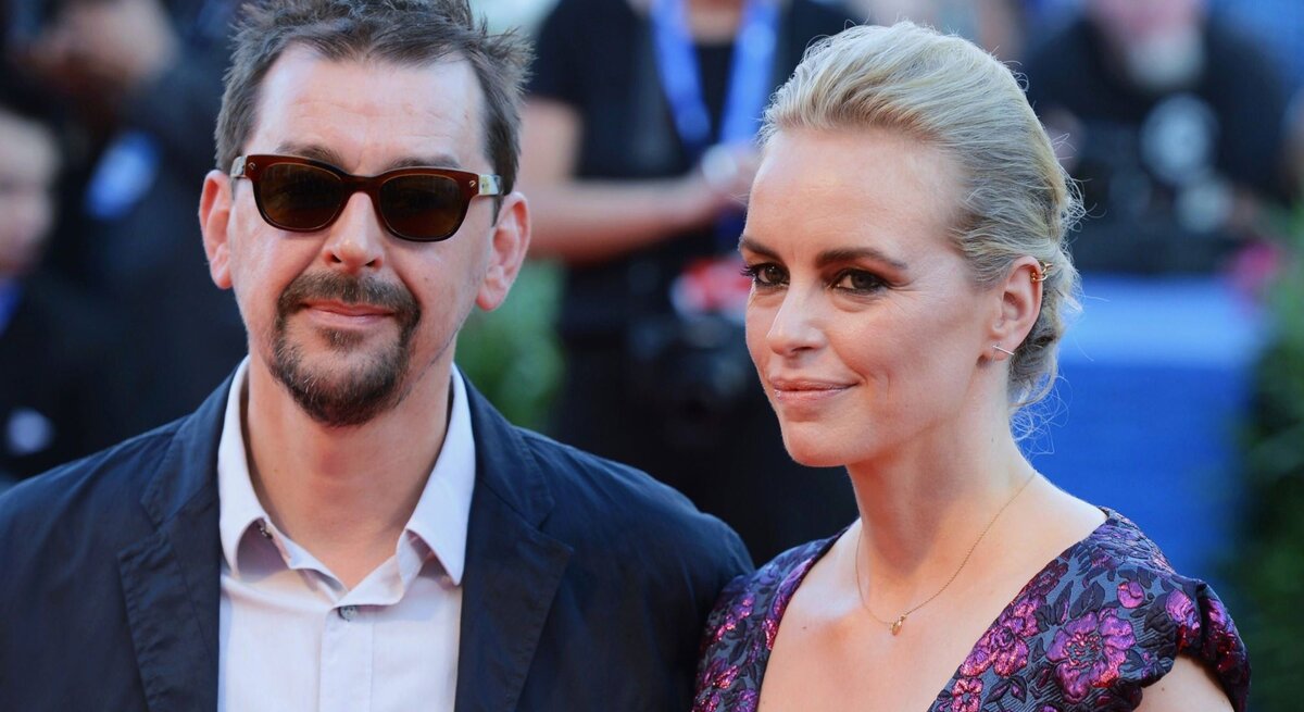 Alex Silva und Nina Hoss stehen Seite an Seite und blicken in die Kamera. Alex Silva trägt eine schwarze Jacke und Sonnenbrille, während Nina Hoss ein Kleid mit Blumenmuster und ein dezentes Make-up trägt. Ihr Haar ist zu einem eleganten Knoten gesteckt. Sie scheinen auf einem roten Teppich zu stehen, erkennbar an den unscharfen Gestalten und Kameras im Hintergrund, was auf eine Film- oder Unterhaltungsveranstaltung hindeutet.
