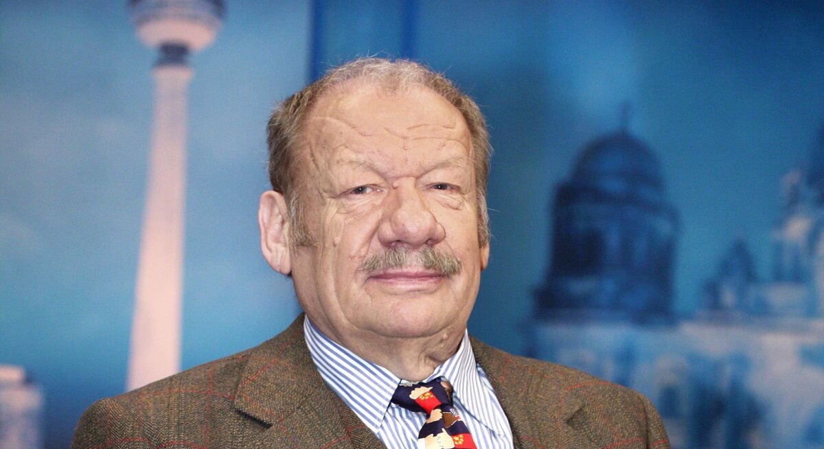 Schauspieler Wolfgang Völz im Anzug schaut ernst in die Kamera, im Hintergrund sind Reichstag und Berliner Fernsehturm als Kulisse zu sehen