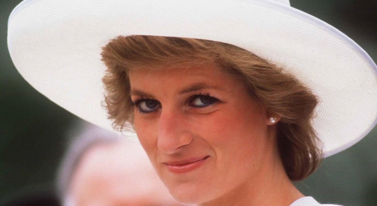 Das Bild zeigt Prinzessin Diana im Jahr 1989 mit ihrem charakteristischen kurzen Haarschnitt, die einen weißen Hut mit breiter Krempe trägt. Sie hat ein freundliches Lächeln und trägt schlichten Schmuck. Prinzessin Diana blickt sanft zur Seite und trägt ein Outfit, das Eleganz und Raffinesse ausstrahlt. Das Bild fängt einen Moment der Ruhe und des Selbstbewusstseins ein, mit einem unscharfen Hintergrund, der die Aufmerksamkeit auf Prinzessin Diana im Vordergrund lenkt.