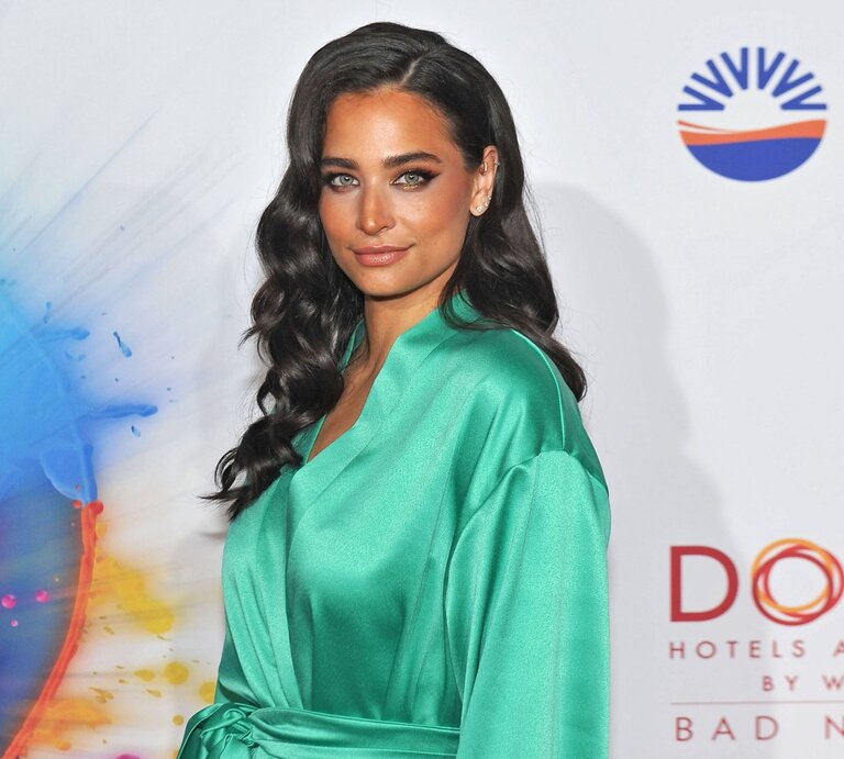 Amira Pocher mit langen, dunklen Locken und einem tiefgrünen Kleid steht vor einer Werbewand. Ihr Blick ist direkt in die Kamera gerichtet und zeigt ein sanftes Lächeln. Ihr Make-up ist elegant mit betonten Augen und natürlich wirkenden Lippen. Die lebendige Farbe ihres Kleides hebt sich gut von der bunten, abstrakten Grafik im Hintergrund ab, die Teil des Werbedisplays zu sein scheint. Sie strahlt Selbstvertrauen und Stil aus. | © IMAGO / STAR-MEDIA