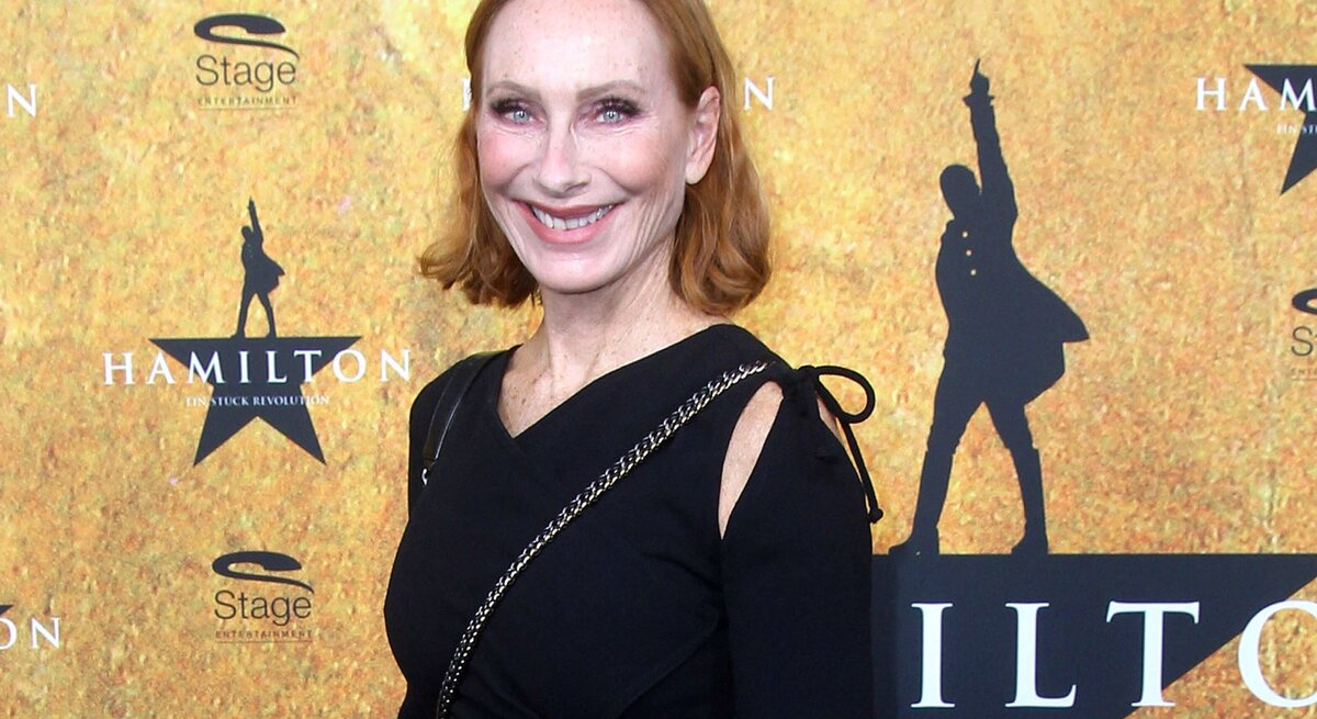 Das Foto zeigt Andrea Sawatzki mit roten Haaren, die lächelnd in die Kamera blickt. Sie trägt ein schwarzes Kleid mit Details an den Ärmeln und steht vor einem Hintergrund, der für das Musical "Hamilton" wirbt. Sie scheint an einer Veranstaltung oder Premiere teilzunehmen, was durch die Logos und den Stern im Hintergrund angedeutet wird.
