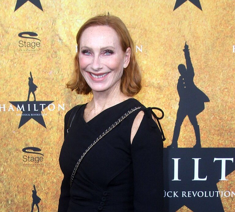 Das Foto zeigt Andrea Sawatzki mit roten Haaren, die lächelnd in die Kamera blickt. Sie trägt ein schwarzes Kleid mit Details an den Ärmeln und steht vor einem Hintergrund, der für das Musical "Hamilton" wirbt. Sie scheint an einer Veranstaltung oder Premiere teilzunehmen, was durch die Logos und den Stern im Hintergrund angedeutet wird. | © IMAGO / APress