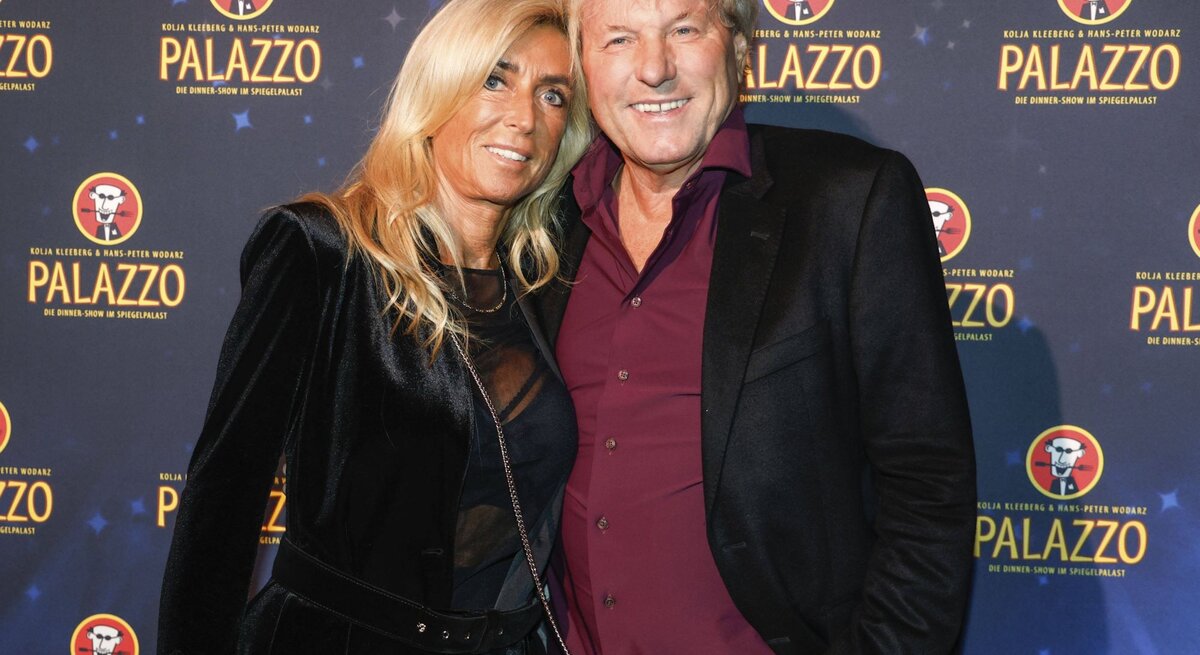 Das Bild zeigt Bernhard Brink und Ute Brink bei einer Veranstaltung auf dem roten Teppich. Hinter ihnen ist ein Hintergrund mit dem Logo "PALAZZO", was darauf hindeutet, dass es sich um eine gesponserte Veranstaltung oder einen Ort handeln könnte, der mit Dinner-Shows oder ähnlichen Unterhaltungsangeboten in Verbindung steht. Sie sind schick gekleidet; Bernhard Brink trägt ein bordeauxrotes Hemd mit einem dunklen Blazer, und Ute Brink trägt eine schwarze Jacke mit passendem Oberteil darunter. Sie scheinen gut gelaunt zu sein und genießen den Anlass.