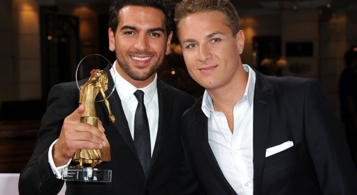 Das Foto zeigt Elyas M'Barek und Joseph M'Barek lächelnd in formeller Kleidung, die einen Preis halten. Elyas M'Barek (links) hält eine Trophäe, die anscheinend bei einer Preisverleihung verliehen wurde. Sie stehen vor einem verschwommenen Hintergrund, der den Anschein eines eleganten Veranstaltungsortes macht. Ihre Kleidung und das Halten der Trophäe deuten darauf hin, dass sie gerade einen Erfolg gefeiert oder eine Auszeichnung erhalten haben.