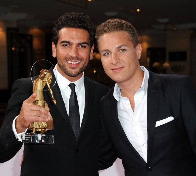 Das Foto zeigt Elyas M'Barek und Joseph M'Barek lächelnd in formeller Kleidung, die einen Preis halten. Elyas M'Barek (links) hält eine Trophäe, die anscheinend bei einer Preisverleihung verliehen wurde. Sie stehen vor einem verschwommenen Hintergrund, der den Anschein eines eleganten Veranstaltungsortes macht. Ihre Kleidung und das Halten der Trophäe deuten darauf hin, dass sie gerade einen Erfolg gefeiert oder eine Auszeichnung erhalten haben. | © IMAGO / Eventpress
