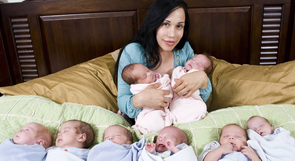 Eine dunkelhaarige Frau in einem blauen Oberteil sitzt auf einem Bett und hält zwei Neugeborene, während sechs weitere Babys in Decken vor ihr liegen.