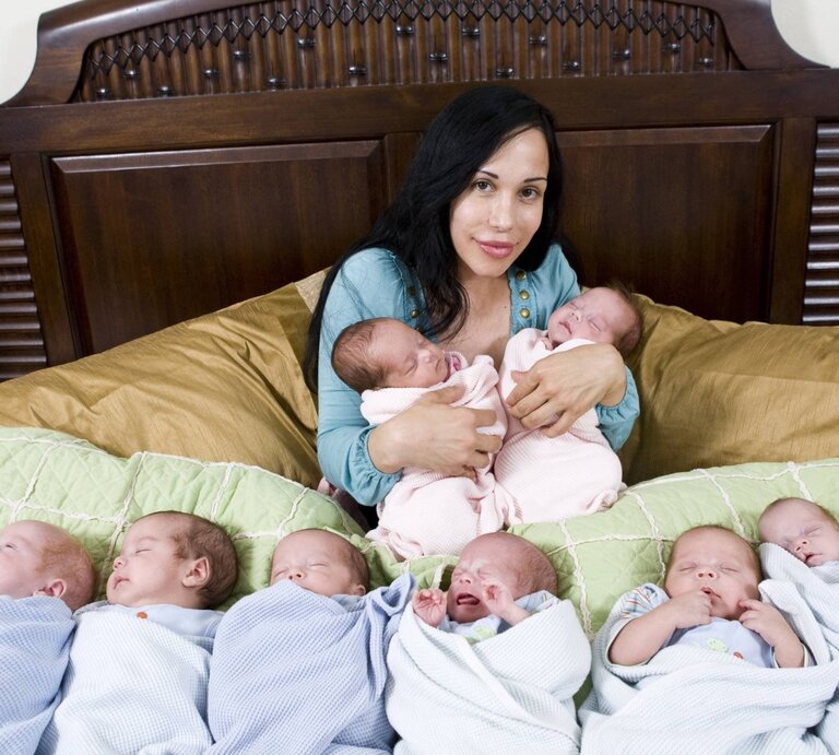 Eine dunkelhaarige Frau in einem blauen Oberteil sitzt auf einem Bett und hält zwei Neugeborene, während sechs weitere Babys in Decken vor ihr liegen. | © IMAGO / Photo News