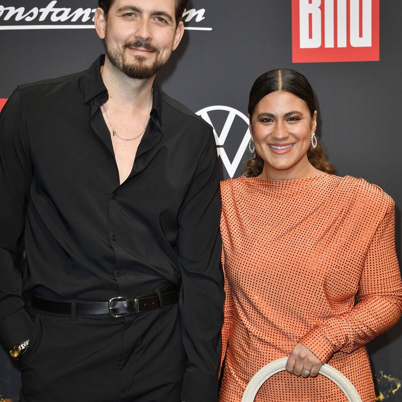 Ein Mann in schwarzem Hemd und Hose hält die Hand einer Frau in einem orangefarbenen Kleid mit Punkten und beigefarbener Tasche. | © IMAGO / Future Image