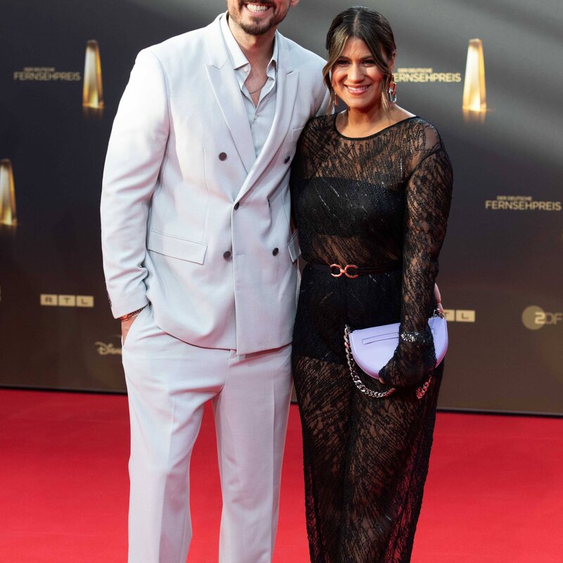 Ein lächelnder Mann im weißen Anzug und eine Frau im schwarzen Spitzenkleid posieren Arm in Arm auf einem roten Teppich. | © IMAGO / Sven Simon