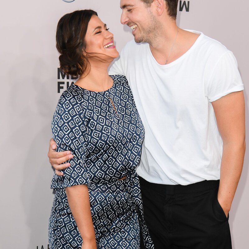 Ein lachender Mann in weißem T-Shirt und schwarzer Hose hält eine Frau im gemusterten Kleid, beide schauen sich liebevoll an. | © Matthias Nareyek/Getty Images for MBFW