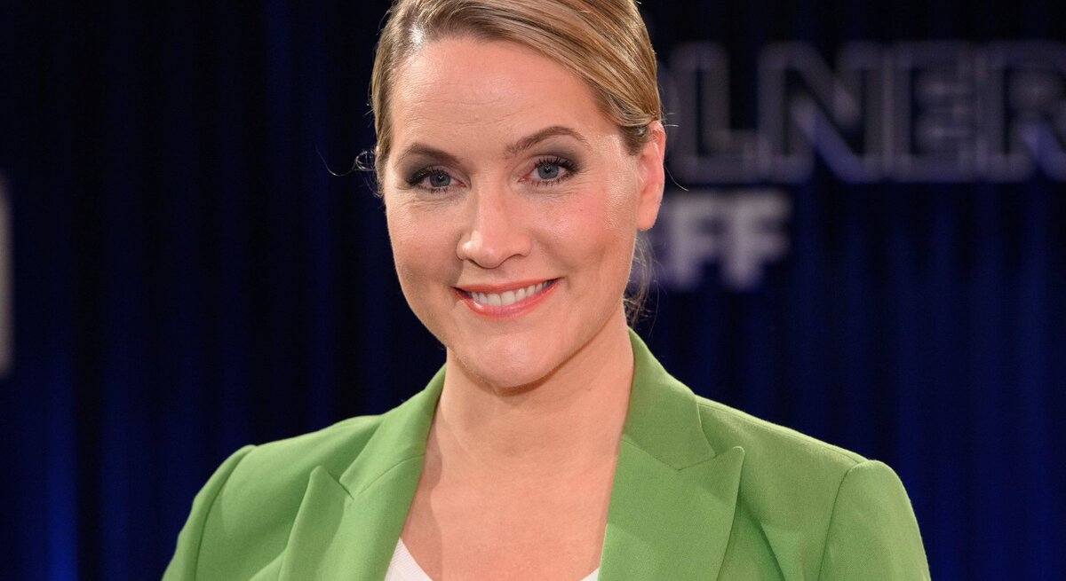 Eine lächelnde Judith Rakers mit zurückgebundenen blonden Haaren, die ein weißes Oberteil und einen leuchtend grünen Blazer trägt, steht vor einem verschwommenen dunkelblauen Hintergrund.
