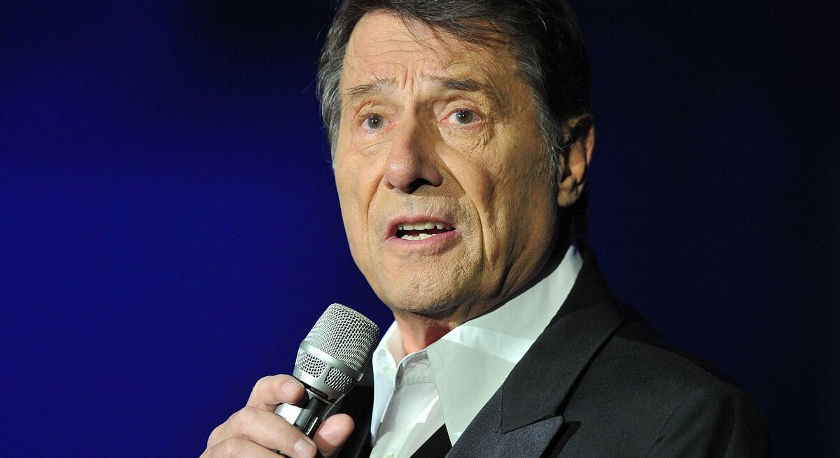 Udo Jürgens trägt einen dunklen Anzug mit rotem Einstecktuch und spricht oder singt in ein Mikrofon vor einem blauen Hintergrund.