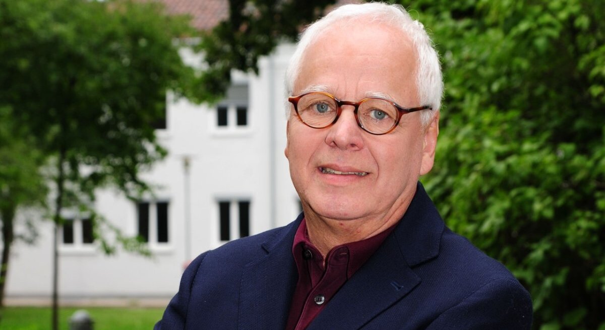 Ein Porträt eines älteren Herren mit grauem Haar und runden Brillen. Er trägt ein dunkelblaues Sakko über einem burgunderroten Hemd. Der Mann blickt direkt in die Kamera und lächelt leicht. Im Hintergrund ist ein unscharfes grünes Laubwerk zu sehen, was auf eine Aufnahme im Freien schließen lässt, mit einem weißen Gebäude im Hintergrund.