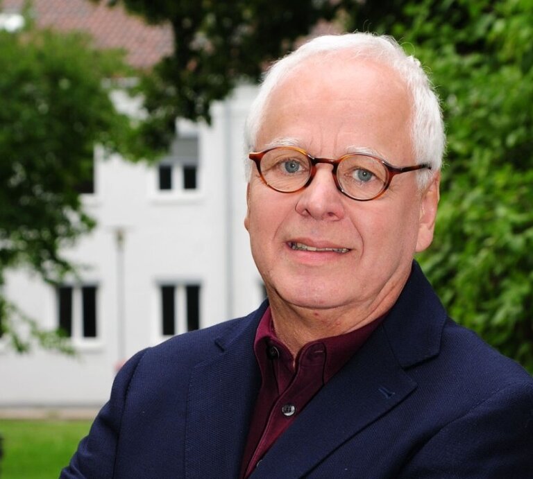 Ein Porträt eines älteren Herren mit grauem Haar und runden Brillen. Er trägt ein dunkelblaues Sakko über einem burgunderroten Hemd. Der Mann blickt direkt in die Kamera und lächelt leicht. Im Hintergrund ist ein unscharfes grünes Laubwerk zu sehen, was auf eine Aufnahme im Freien schließen lässt, mit einem weißen Gebäude im Hintergrund. | © Peter Bischoff/Getty Images