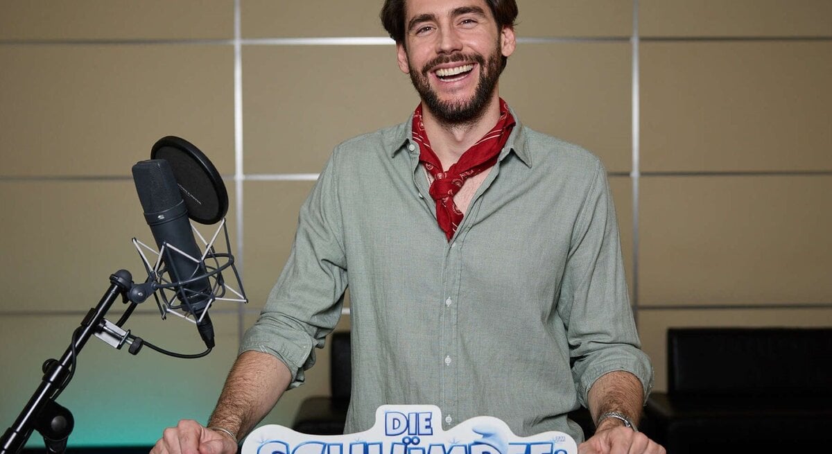 Alvaro Soler mit braunem Bart und grünem Hemd steht vor Mikrofon, während er für "Die Schlümpfe" Werbung macht.