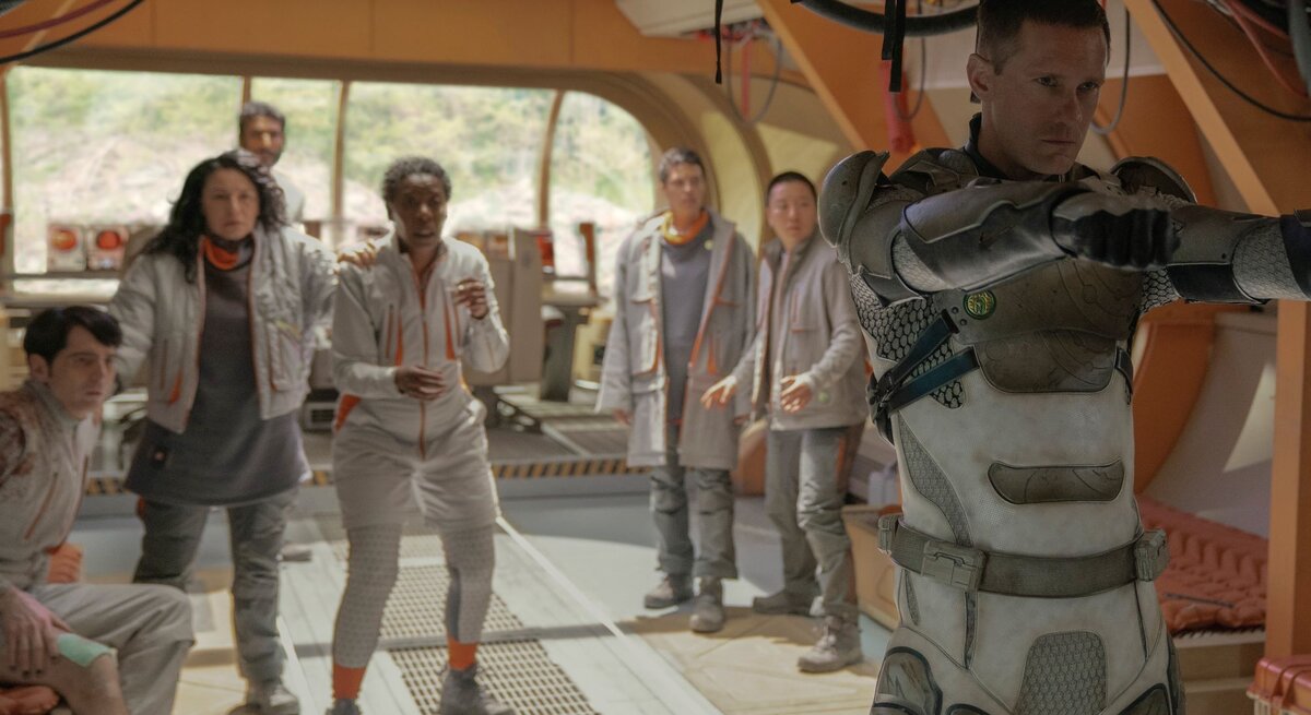 Alexander Skarsgard in "Murderbot": Ein ernster Mann in futuristischer Rüstung zielt mit erhobenen Armen, während sieben verängstigte Personen hinter ihm stehen.