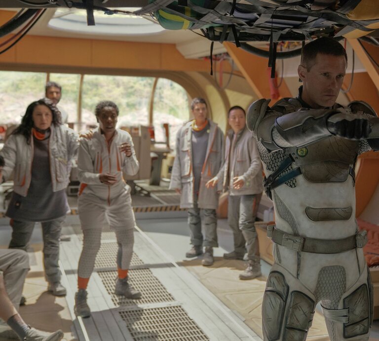 Alexander Skarsgard in "Murderbot": Ein ernster Mann in futuristischer Rüstung zielt mit erhobenen Armen, während sieben verängstigte Personen hinter ihm stehen. | © Apple TV+