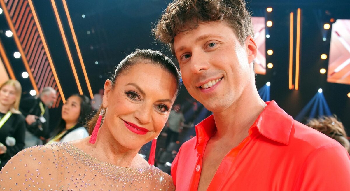 Christine Neubauer im glitzernden Kleid mit langen pinken Ohrringen und Valentin Lusin in offenem roten Hemd lächeln in die Kamera.
