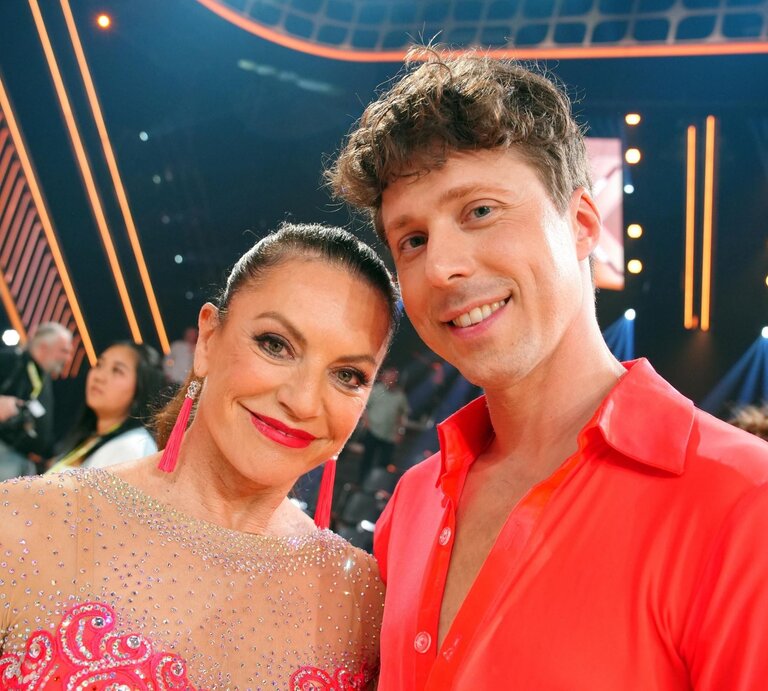 Christine Neubauer im glitzernden Kleid mit langen pinken Ohrringen und Valentin Lusin in offenem roten Hemd lächeln in die Kamera. | © RTL / Stefan Gregorowius