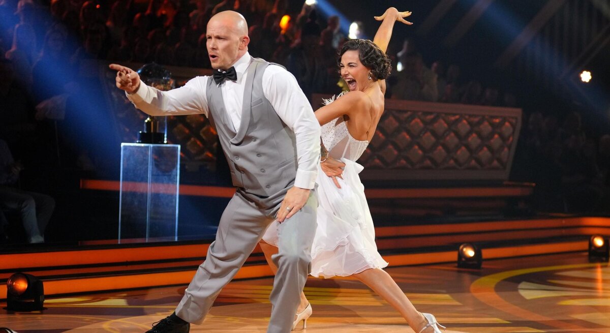 Fabian Hambüchen im grauen Anzug tanzt energiegeladen mit lachender Anastasia Maruster in weißem Kleid auf beleuchteter Showbühne.