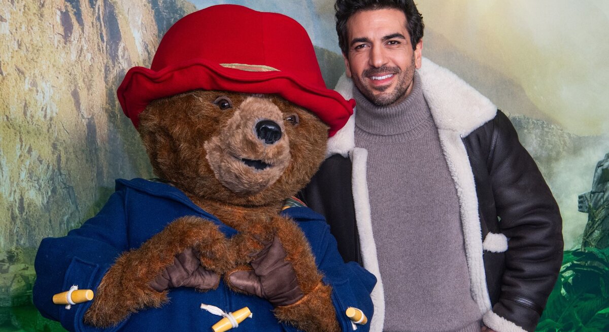 Elyas M'Barek in brauner Lederjacke mit Fellkragen posiert neben Paddington Bär, der eine rote Mütze und blauen Mantel trägt.