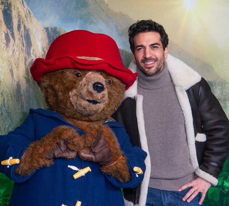 Elyas M'Barek in brauner Lederjacke mit Fellkragen posiert neben Paddington Bär, der eine rote Mütze und blauen Mantel trägt. | © Matthias Nareyek/Getty Images