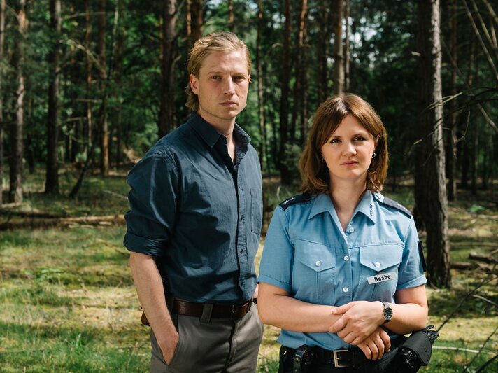 Ein blonder Mann in dunkelblauem Hemd und eine Polizistin mit braunen Haaren stehen ernst blickend in einem sonnigen Wald. | © rbb/ Elliott Kreyenberg
